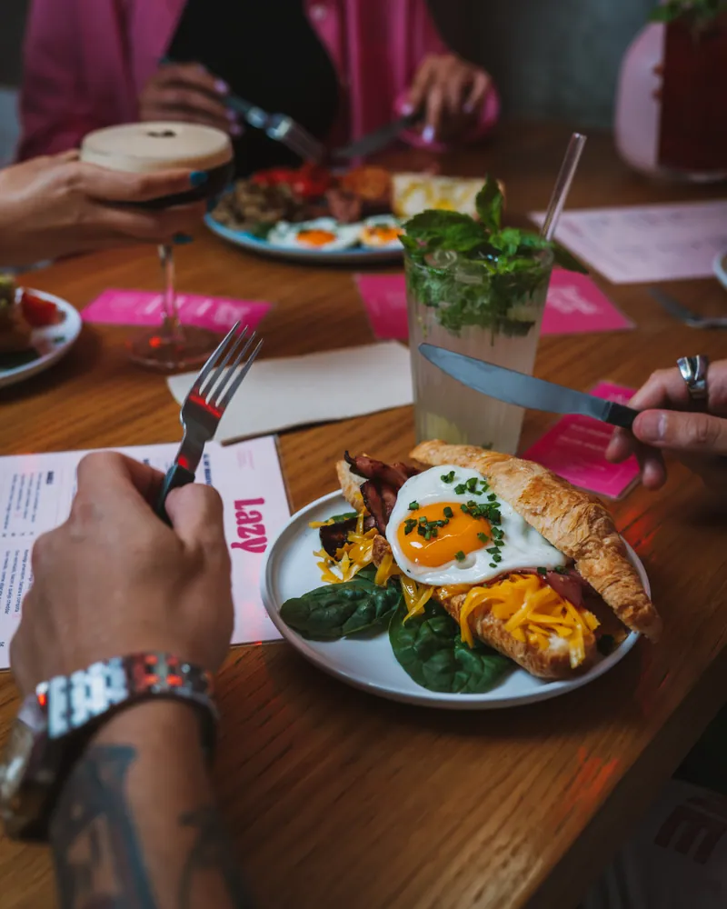 Croissant com ovo estrelado, bacon e cheddar numa mesa cheia de gente